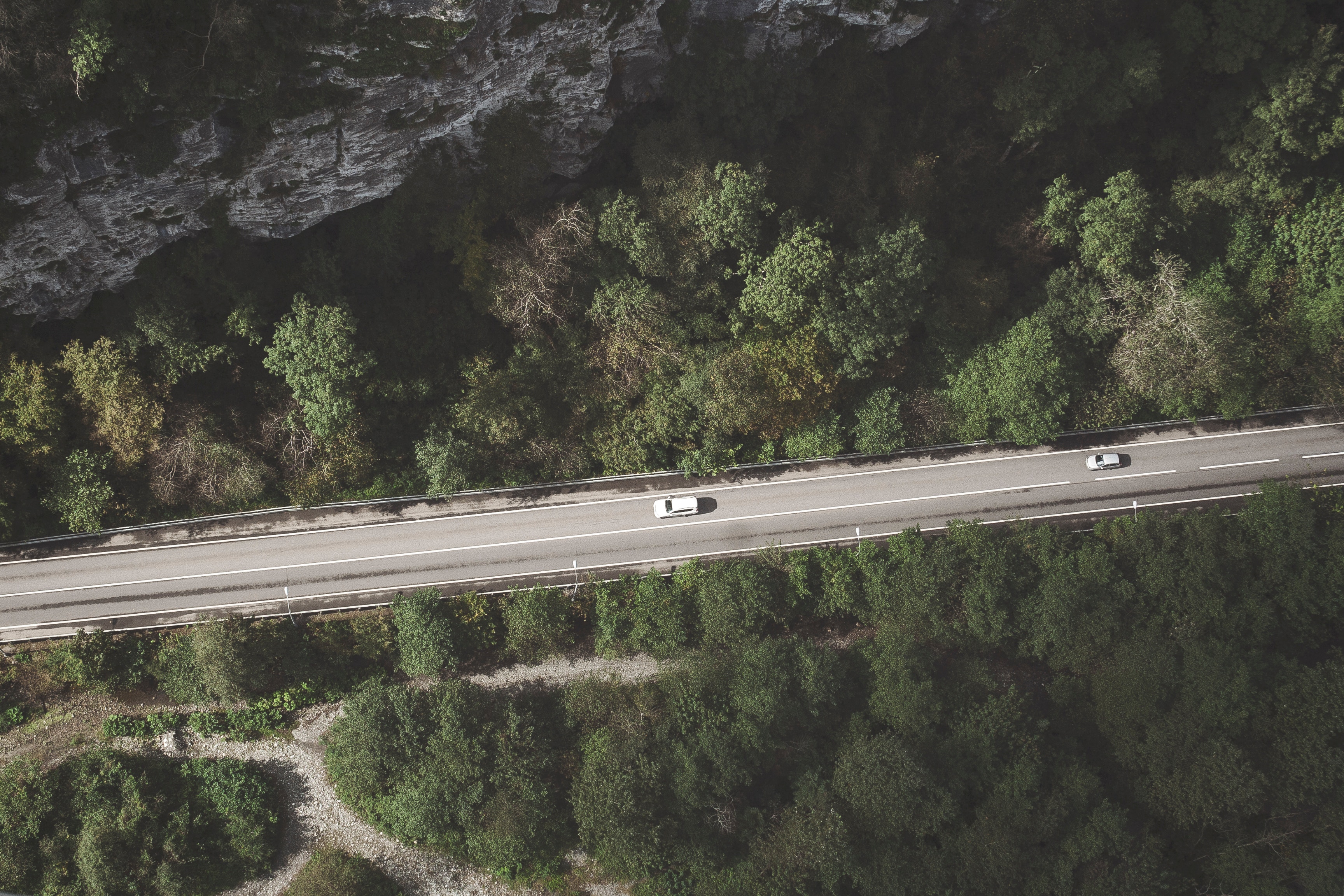 En vit bil kör på en landsväg omgiven av skog, med en framträdande klippformation till höger.