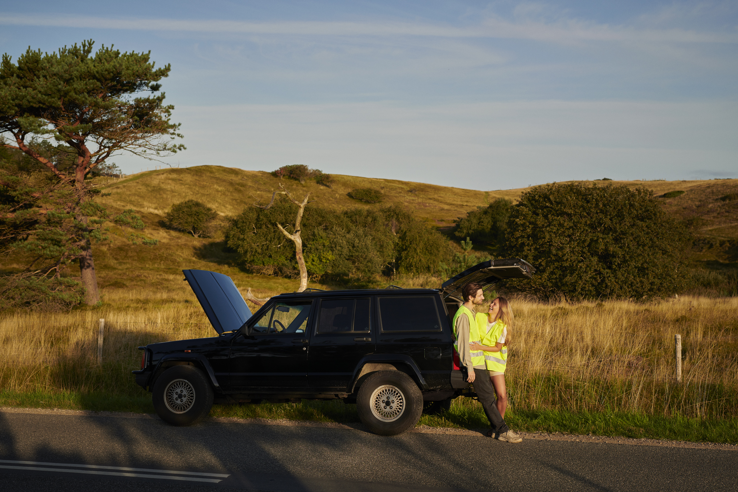 En man och en kvinna, klädda i gulfärgade västar, sitter och skrattar i bakluckan på en parkerad svart jeep längs en landsväg.