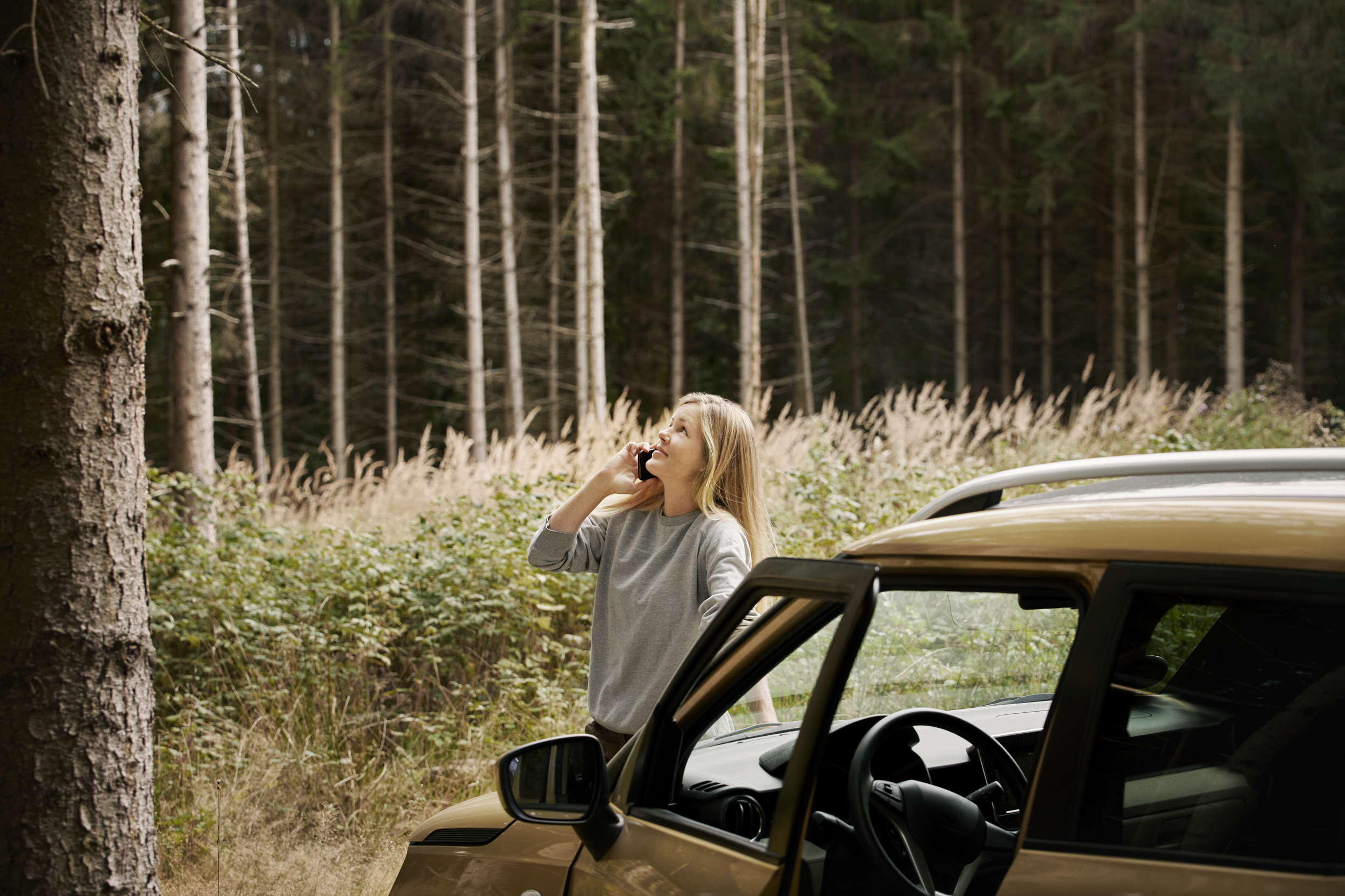En kvinna står vid sin parkerade guldfärgade bil i skogen, pratar i telefon och tittar upp mot trädet framför henne.