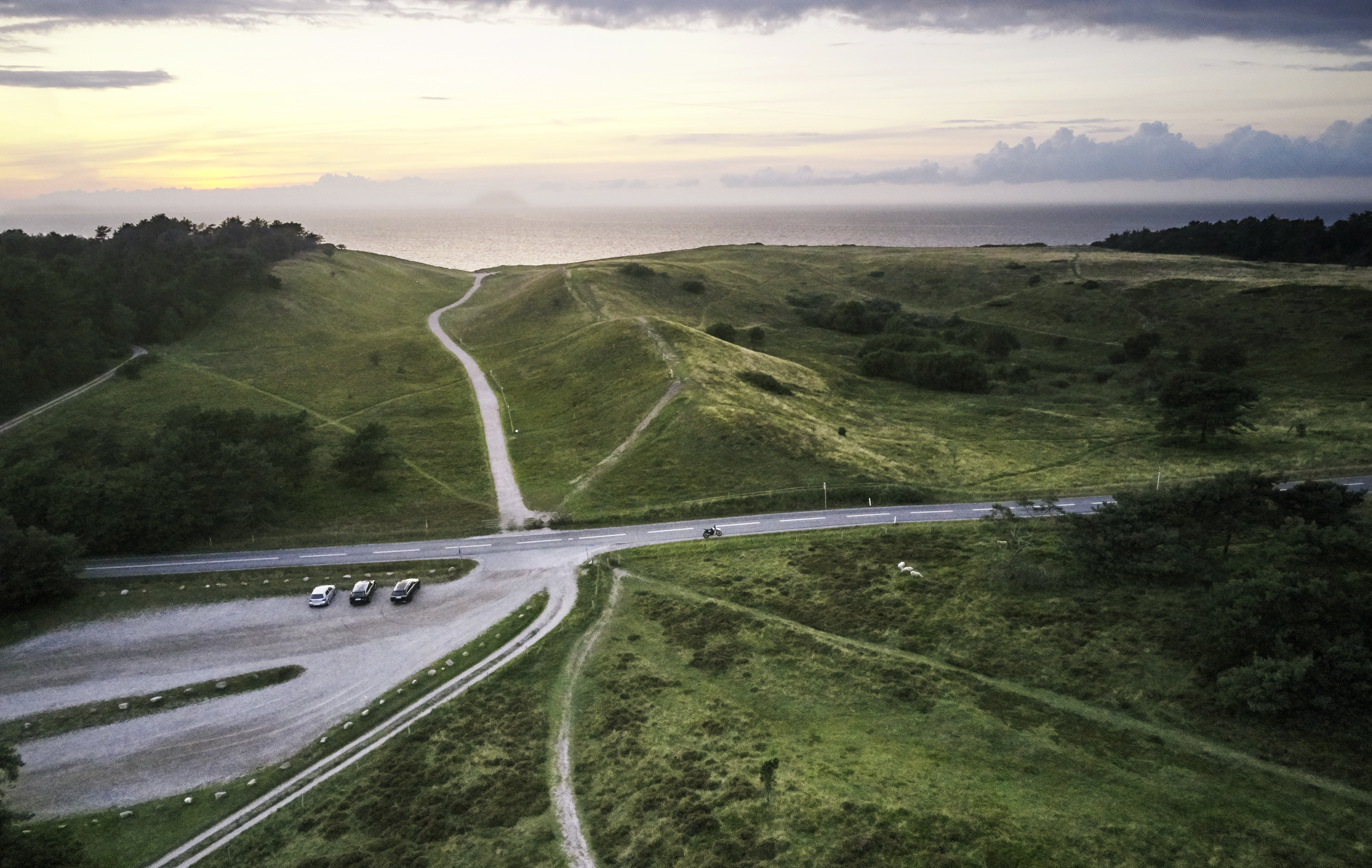 En landsväg som slingrar sig genom ett grönt landskap med en parkeringsplats bredvid, där tre bilar står parkerade.