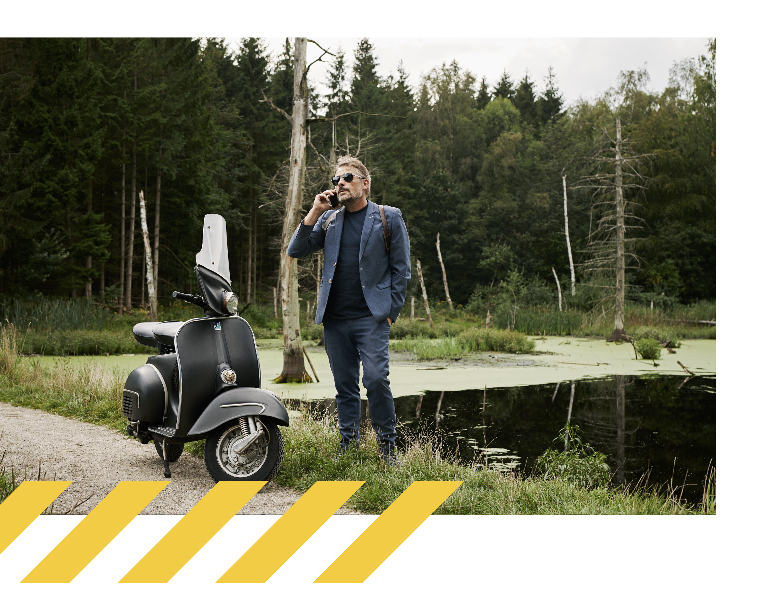 En man i blå kavaj står vid sidan av en väg vid en sjö, pratar i telefon bredvid sin parkerade svarta moped.