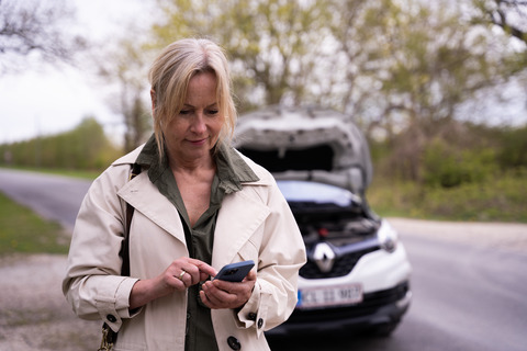 En kvinna som står framför sin bil med framluckan öppen och använder sin telefon.