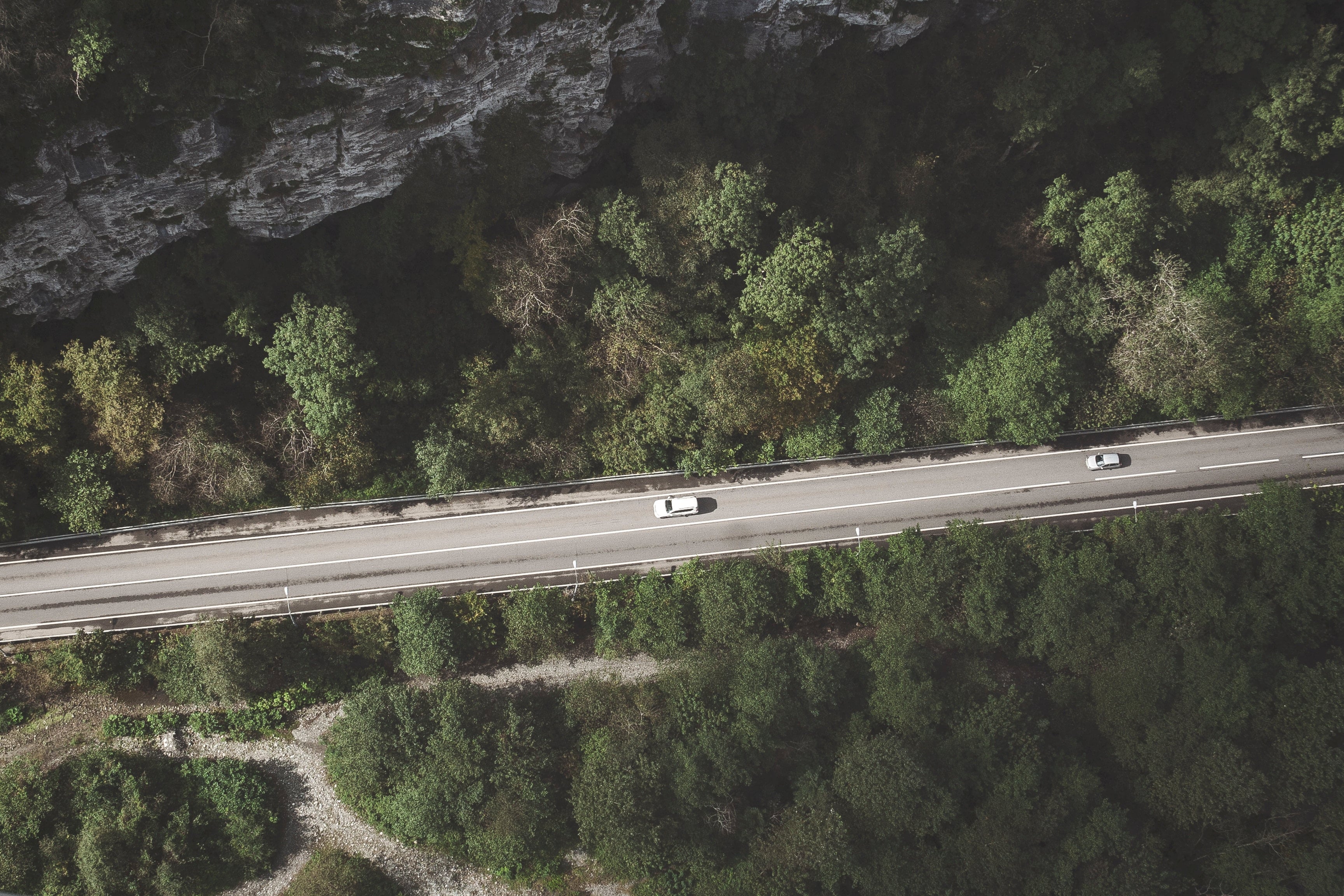 En vit bil följs av en silverfärgad bil längs en landsväg omgiven av skog, med en framträdande klippformation till höger.