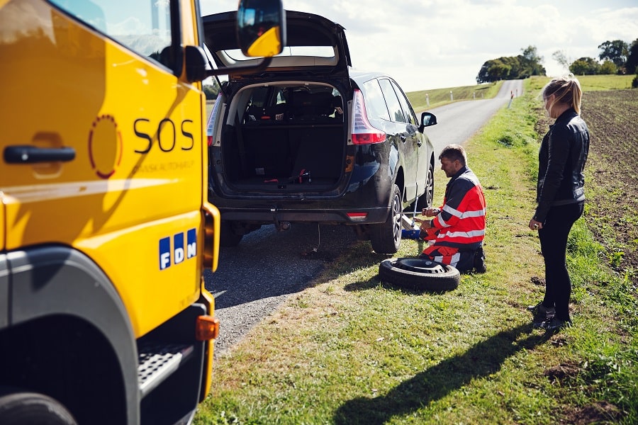 En bärgningsarbetare assisterar en kvinna i svart med att byta däck på hennes svarta bil vid sidan av en landsväg. Bärgningsfordonet är parkerat bakom den svarta bilen.