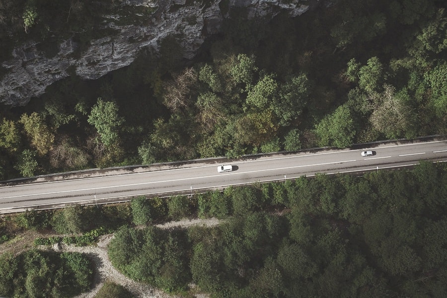En vit bil följs av en silverfärgad bil längs en landsväg omgiven av skog, med en framträdande klippformation till höger.