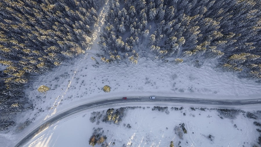 En röd bil och en silverfärgad bil färdas längs en landsväg som slingrar sig genom ett vinterlandskap, rikt dekorerat med snötäckta granar.